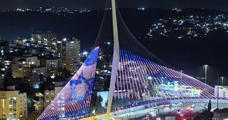 גשר המיתרים בירושלים מואר בלילה בצבעי כחול-אדום, עם הדמיית דגל ישראל