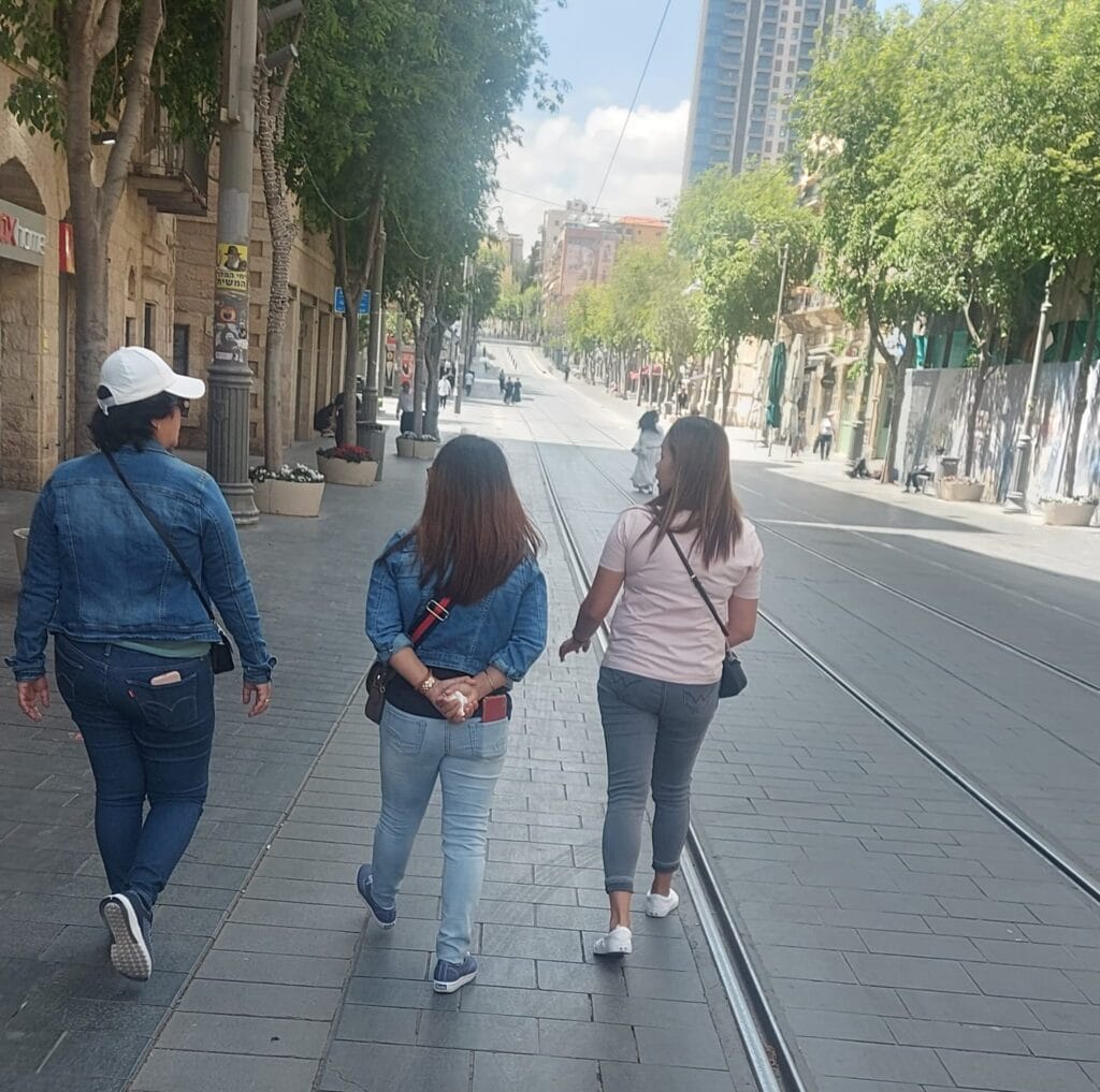 רחוב יפו בירושלים ביום שמש, נשים צועדות לצד מסלול הרכבת הקלה, עצים ירוקים ובניינים עירוניים ברקע