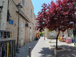 רחוב משעול מסילת ישרים בירושלים עם עץ פורח, דוכנים וסמטה שקטה ביום שמשי