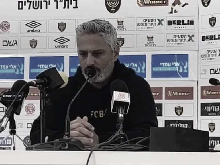 Barak Itzhaki, head coach of Beitar Yerushalayim, at a press conference, looking serious and silent behind a row of microphones